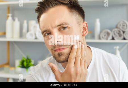Man in bathroom applying cream to his face, touching cheek and looking intently, self care grooming Stock Photo