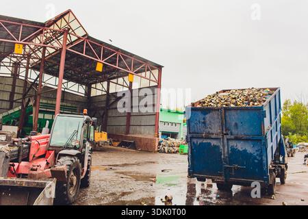 Ufa, Russia - 13 June 2025 Open yard with truck and tractor, container filled with recyclables, muddy ground with puddles, canopy steel frames Stock Photo