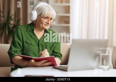 Elderly woman taking notes while wearing headphones and using laptop at home Stock Photo