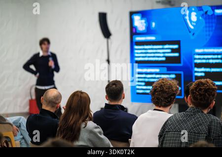 Speaker presents to attentive group in bright studio style room. Large screen shows blue graphics and text Audience seated listening taking notes. Collaborative learning atmosphere modern presentation Stock Photo