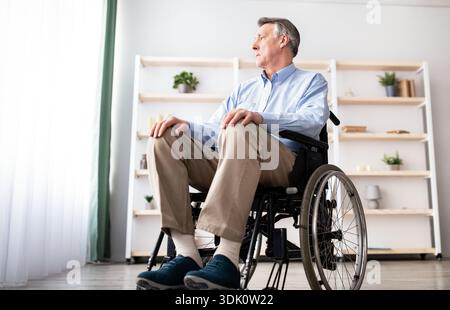 Older Man Using a Wheelchair Sits in a Bright Room and Looks out the Window Stock Photo