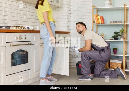 Man plumber fixing broken pipe in modern kitchen and talking with female client at home. Stock Photo