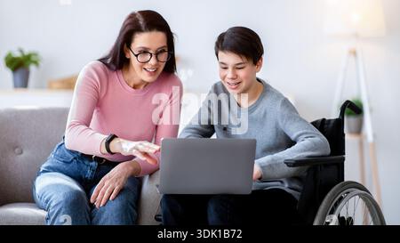 Woman With Glasses Helps Boy Using Wheelchair With Laptop in a Modern Living Room Stock Photo