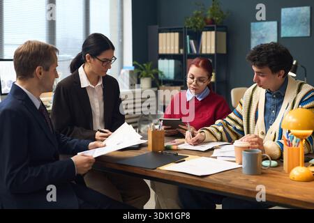 Group of young adult and middle aged multiethnic colleagues collaborating at office table, reviewing documents and using digital tablet, focused on teamwork and project discussion Stock Photo