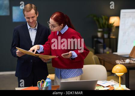 Caucasian middle aged man and Caucasian young adult woman reviewing documents together in modern office setting, woman holding folder and gesturing while man listening attentively Stock Photo