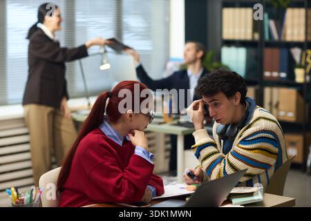 Young Caucasian woman and young Caucasian man sitting at desk discussing work, man holding pen and looking concerned, two middle aged colleagues working in background in modern office Stock Photo