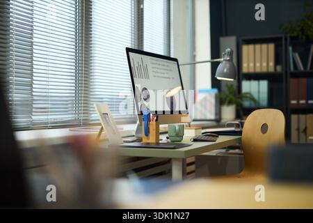 Modern office workspace featuring desktop computer displaying business charts and graphs, organized desk with stationery, coffee mug, and window blinds allowing natural light Stock Photo