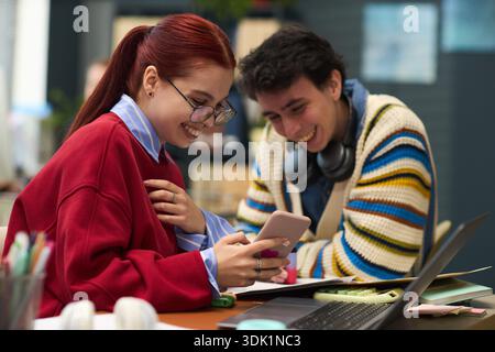 Young Caucasian woman and young Caucasian man sitting at desk smiling and looking at smartphone together, engaging in conversation, studying or working in modern office setting Stock Photo