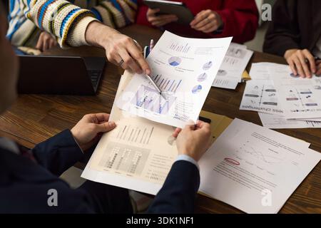 Caucasian middle aged man holding financial charts while multiethnic group of young adults discussing business strategy around table with printed graphs and digital tablet Stock Photo