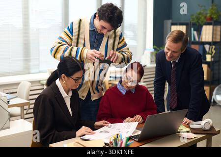 Company employees including Caucasian male executive, senior female manager, two young trainees collaborating in office, teenager photographing documents with smartphone while discussing project tasks Stock Photo