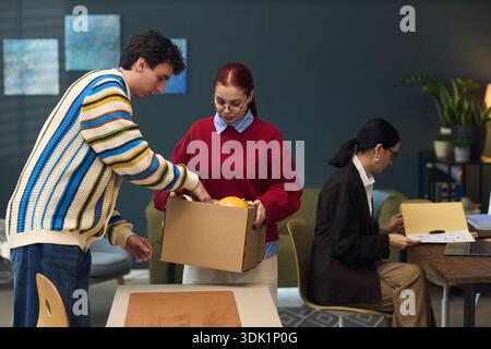 Caucasian young man assisting Caucasian young woman holding cardboard box while female manager aged woman working on laptop at desk in modern office setting Stock Photo