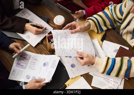Group of young adult and middle aged Caucasian and Black hands analyzing business charts and graphs at table, holding printed documents with data, discussing financial strategy, coffee cup visible Stock Photo