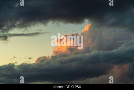 Dramatic evening sky with one cloud catching warm sunset light above dark storm background. Natural weather concept emphasizing light versus darkness. Stock Photo