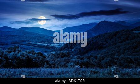 mountainous countryside scenery surrounded with forested rolling hills at night. rural area peaceful landscape with dark sky in full moon light. early Stock Photo