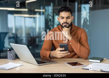 Young indian man in a contemporary office setting sitting at a wooden desk with a laptop, tablet, and documents, carefully reviewing information on hi Stock Photo