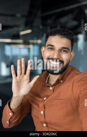 Friendly young man looking at camera and waving his hand, greeting or saying goodbye during an online meeting or taking a cheerful selfie in a contemp Stock Photo