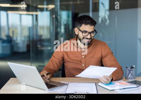 Young businessman working in a bright modern office, smiling while reviewing documents and using a laptop, representing productivity, success, and a p Stock Photo