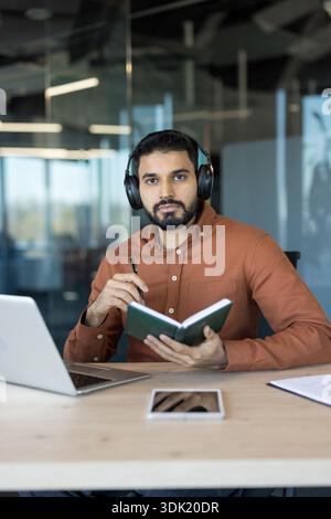Young man wearing headphones and concentrating on work, holding a notebook and pen while sitting at a desk with a laptop and tablet, engaged in remote Stock Photo
