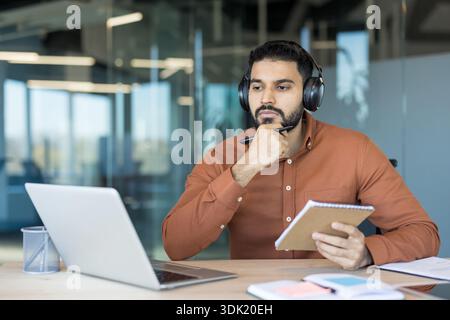 Young man wearing headphones and holding a pen, listening attentively and taking notes on a notepad while working on a laptop in a modern office or at Stock Photo