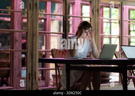 Tired or bored caucasian female worker or student sit at desk need rest suffer from fatigue overworked at public place. Exhausted woman working at computer, feeling headache, try to do her project || Model released Stock Photo