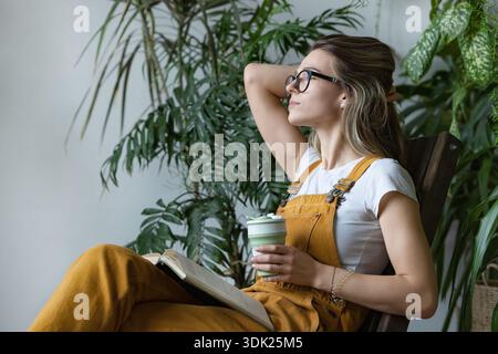 Woman gardener wearing overalls, resting after work, sitting on wooden chair in home greenhouse, hold reusable coffee or tea mug, looking at the window with a open book on knees. Break during work || Model released Stock Photo