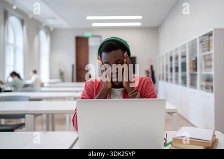 Education and mental health. Stressed overwhelmed african american guy student tired of learning sitting in college library holding head in hands and looking at laptop with frustrated face expression || Model released Stock Photo