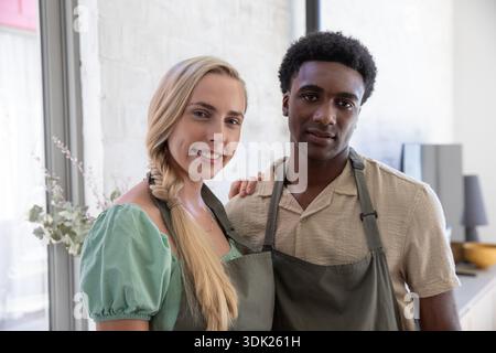 Diverse couple wearing grey aprons standing in modern kitchen near glass vase and wooden bowl. Cooking, collaboration, minimalism, lifestyle, interior, wellness, home || Model released Stock Photo