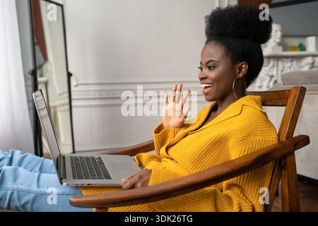 Smiling African American biracial millennial woman with afro hairstyle in yellow cardigan resting, sitting on armchair, talking in video chat with her friends, say hello. || Model released Stock Photo
