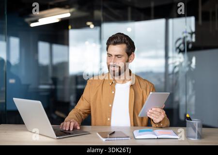 Person working on a laptop and holding a digital tablet, navigating multiple screens for efficient remote work and productivity in a contemporary busi Stock Photo
