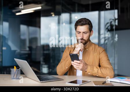 Bearded professional man deep in thought browsing content on his smartphone at a modern office desk with laptop, tablet and notebook, planning and wor Stock Photo