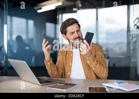 Stressed businessman in a modern office having an emotional outburst while holding a mobile phone, expressing frustration and anger at an online probl Stock Photo