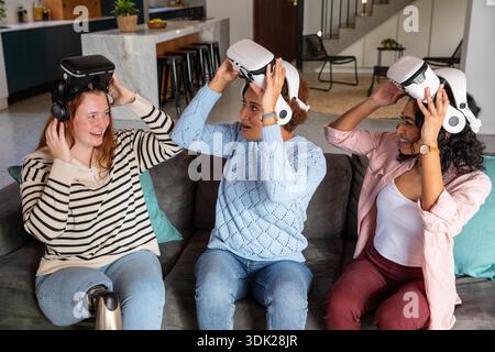 Diverse female friends sitting on sofa lifting white VR headsets in living room near marble island. Gaming, entertainment, relaxation, modern, home, leisure, technology || Model released Stock Photo