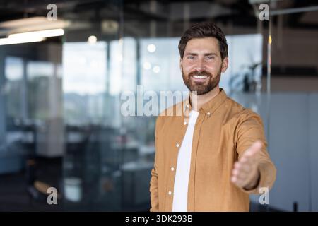 Young businessman standing in a modern office, smiling confidently and offering a welcoming handshake, representing new opportunities, trust, and succ Stock Photo