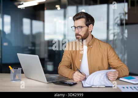 Young man in glasses focusing on financial data and reports, analyzing numbers and managing business tasks at a modern office desk, utilizing a laptop Stock Photo