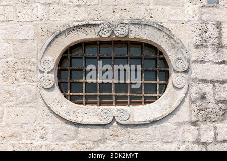 A rustic oval window set in a light stone wall, protected by worn iron bars and framed by decorative stonework. This architectural detail evokes histo Stock Photo