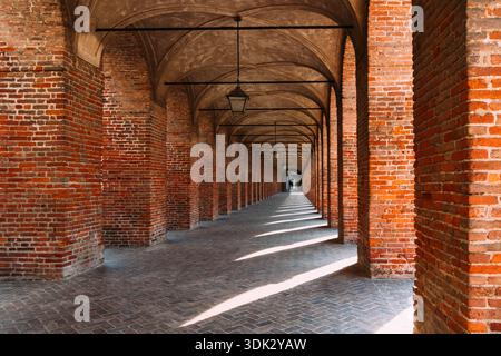 The image captures a long, brick arcade in Sabbioneta, Italy, with a symmetrical design and repeating arches. Sunlight creates shadows. Stock Photo