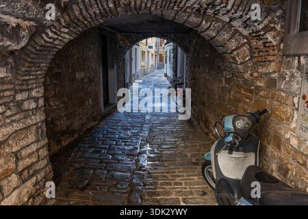 Stone wall passage under apartment building with scooter and bike on cobblestone street in medieval Old Town of Piran in  Slovenia. Stock Photo