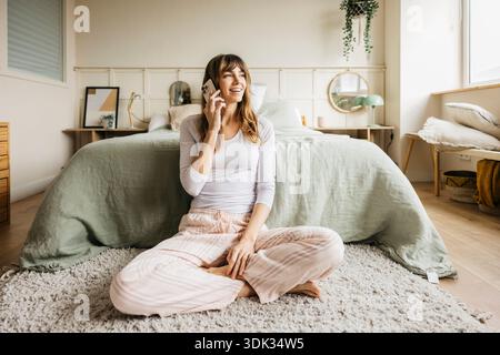Woman in pajamas talking on her mobile phone while sitting on the floor in her modern bedroom Stock Photo