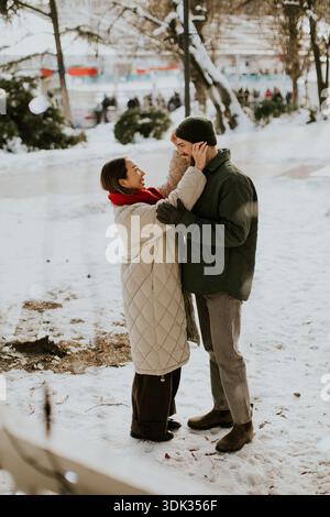A Korean woman and a Caucasian man stand in a snowy park. They smile at each other while the woman touches the man's face. Trees and people are visibl Stock Photo