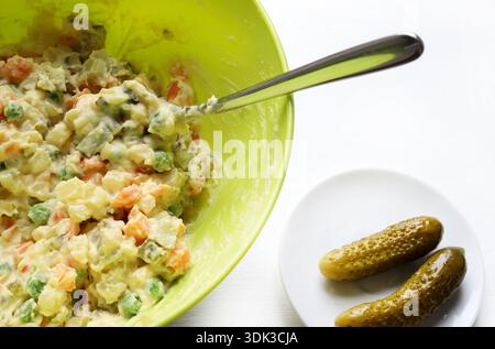 Olivier salad in a green bowl with a spoon on a white background. Cold appetizer with boiled vegetables, pickled cucumbers, and mayonnaise. Stock Photo