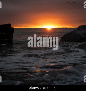 Dramatic sunset over the ocean with silhouetted pier ruins, large rocks, crashing waves, and a seagull against fiery orange skies Stock Photo