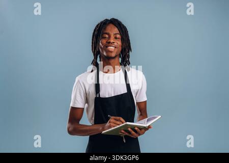 Standing, barista is in apron. Black man is in the studio against soft background. Stock Photo