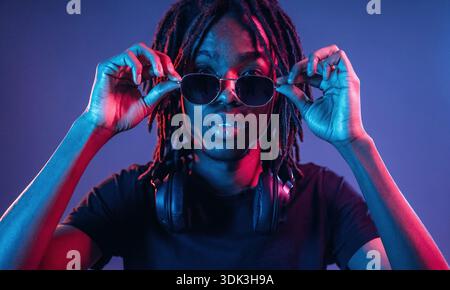 Touching sunglasses, front view. African American guy is studio with neon lighting. Stock Photo