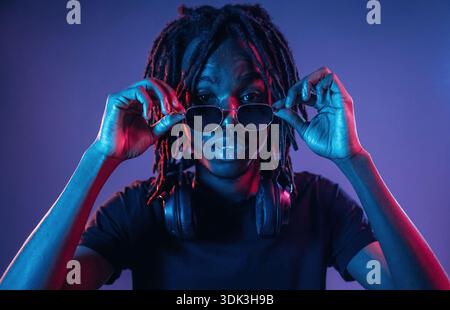 Touching sunglasses, front view. African American guy is studio with neon lighting. Stock Photo