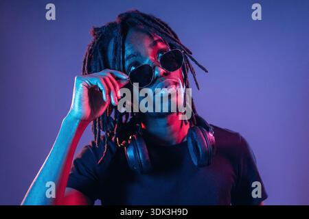 Touching sunglasses, front view. African American guy is studio with neon lighting. Stock Photo