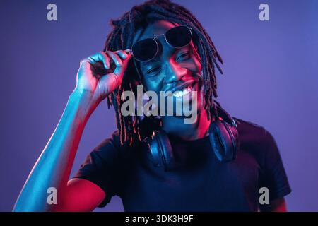 Touching sunglasses, front view. African American guy is studio with neon lighting. Stock Photo