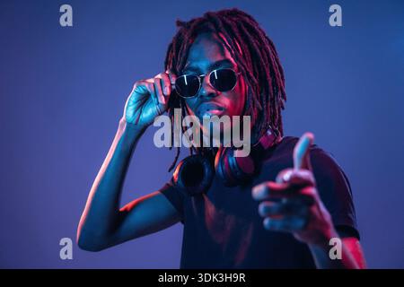 Pointing by finger, in sunglasses. African American guy is studio with neon lighting. Stock Photo