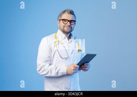 Male doctor in white coat with stethoscope uses a tablet on blue studio background. Professional medical concept for digital healthcare services, onli Stock Photo