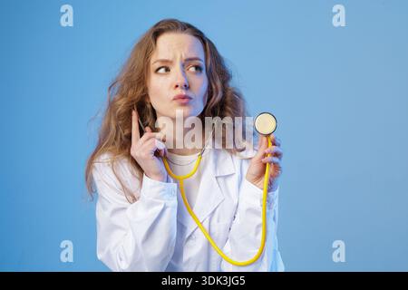 Female doctor in white coat holds a stethoscope and looks aside thoughtfully in studio. Clean medical concept for healthcare diagnosis awareness, educ Stock Photo