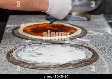 Pizzaiolo spreading red tomato sauce on raw dough base with spoon during pizza preparation process. Stock Photo